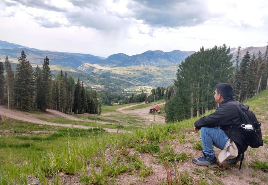 Gino Di Angelo Machoa Flores, sentado en una ladera, observa un valle con bosques de coníferas y montañas en Colorado, Estados Unidos
